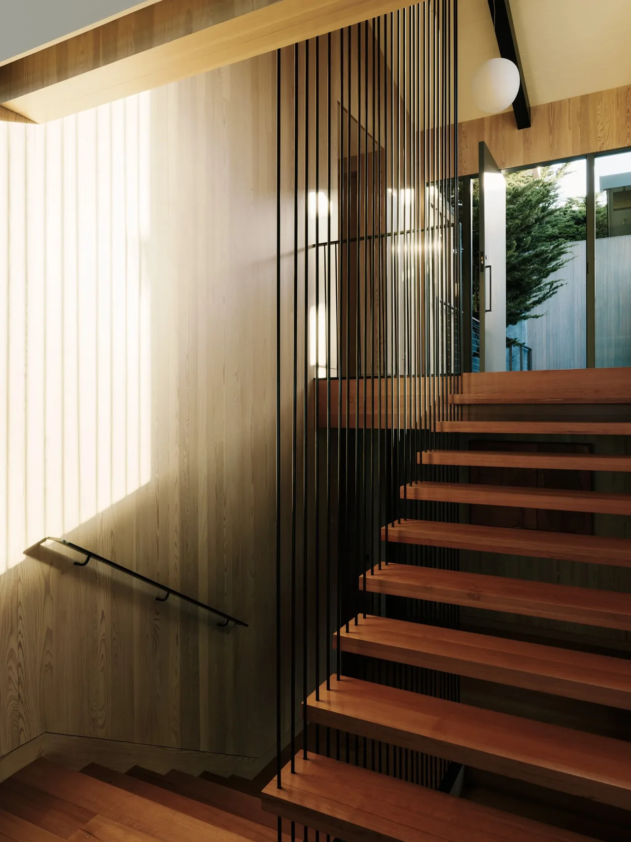 Floating wood stairs with vertical black metal railings in a wood-paneled room with a glass entry door.