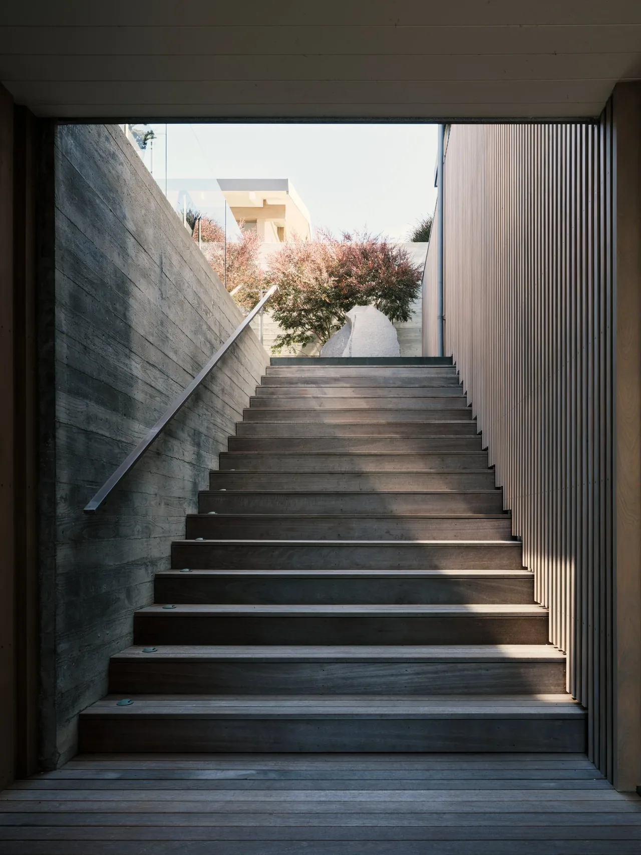 Outdoor staircase framed by a concrete wall on one side and vertical wood paneling on the other, leading to an upper garden space.