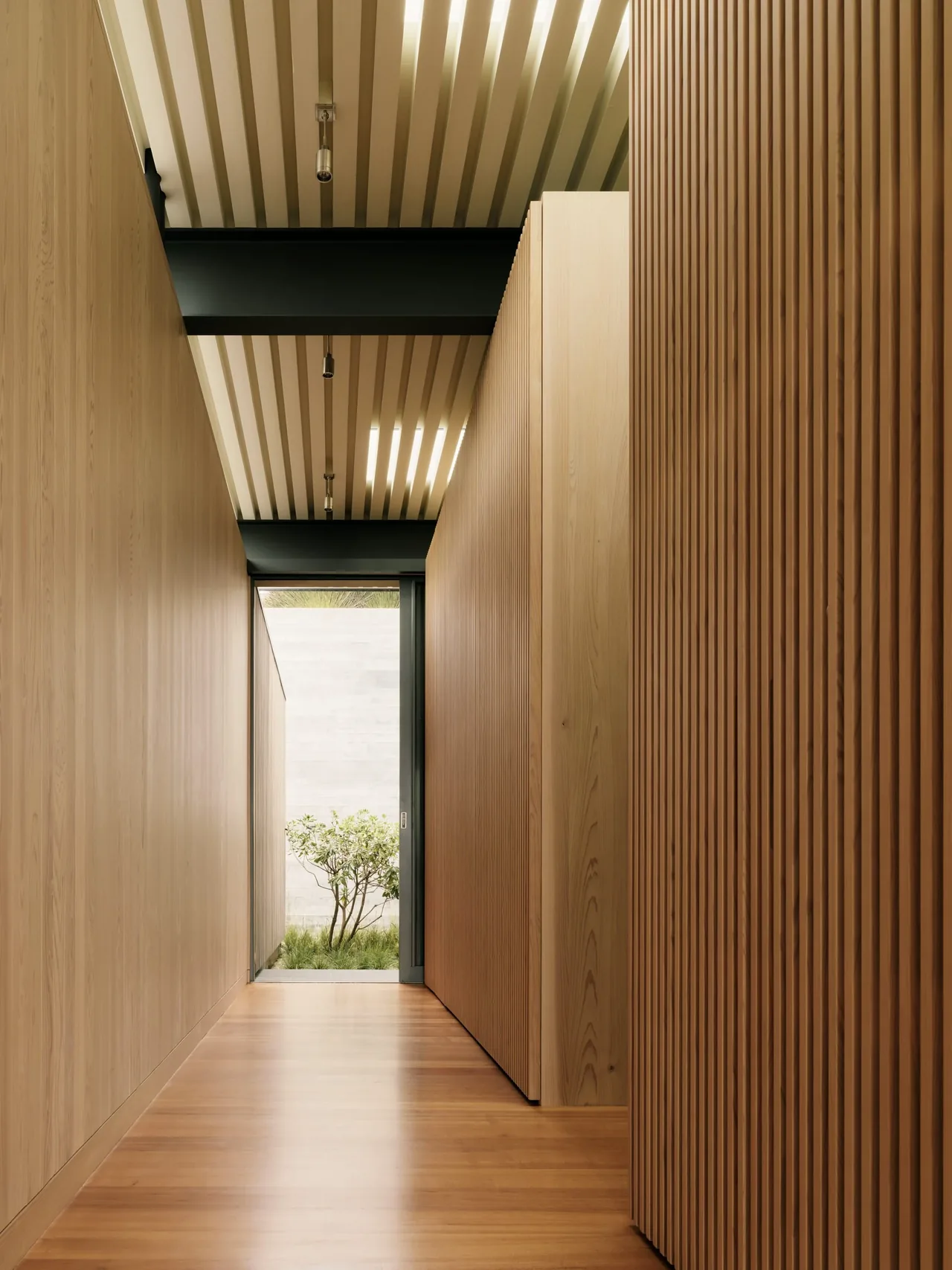 The hallway features smooth wood paneling, vertical wood slats on one wall, and exposed ceiling beams leading to a partially visible garden outside.