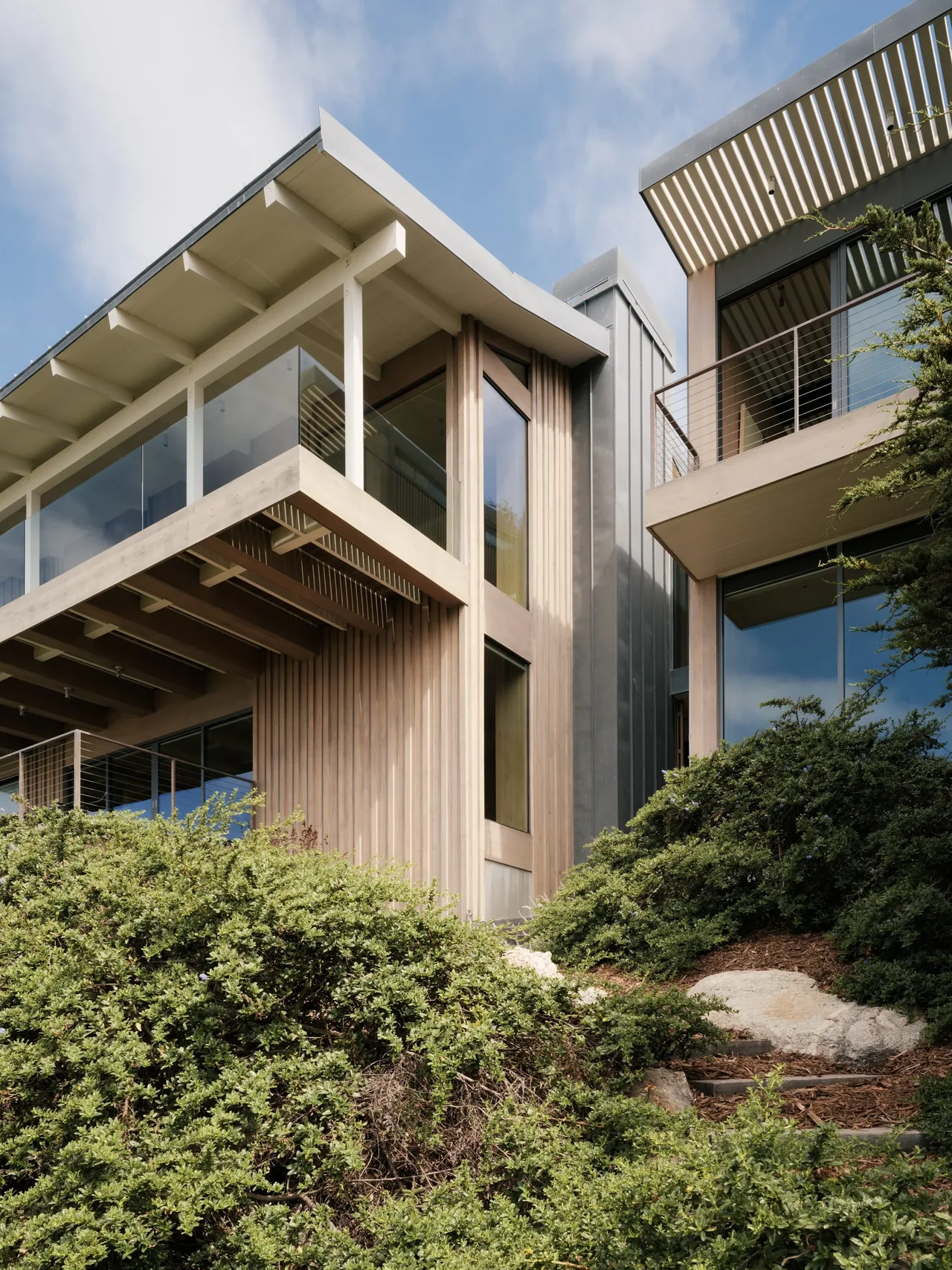 Modern house exterior with vertical wood slat siding, glass railings on balconies, and metal panel siding.