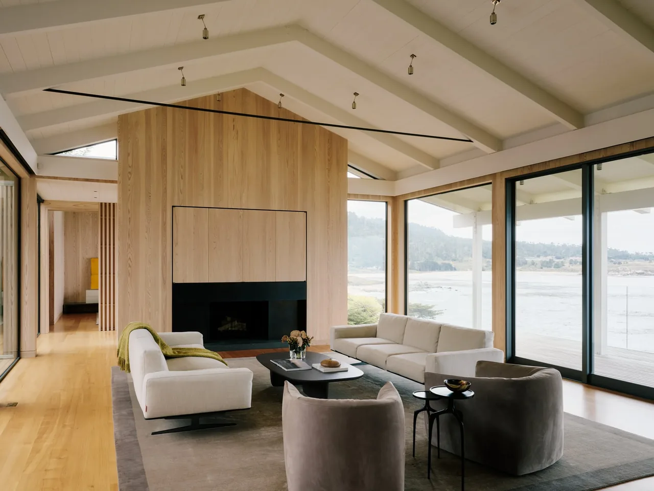 Living room with vaulted ceiling, wood-paneled fireplace, and large floor-to-ceiling windows framing a coastal landscape.