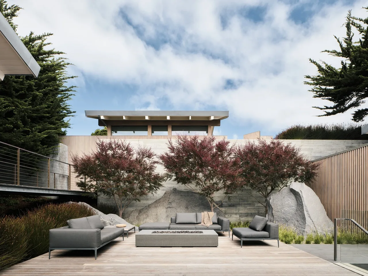Modern outdoor seating space in a coastal residential setting with a rectangular fire pit and three trees.