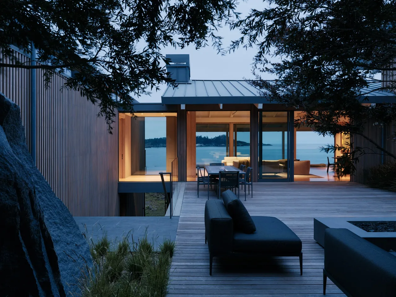 Modern home exterior with a wood deck and glass walls offering a water view at dusk.