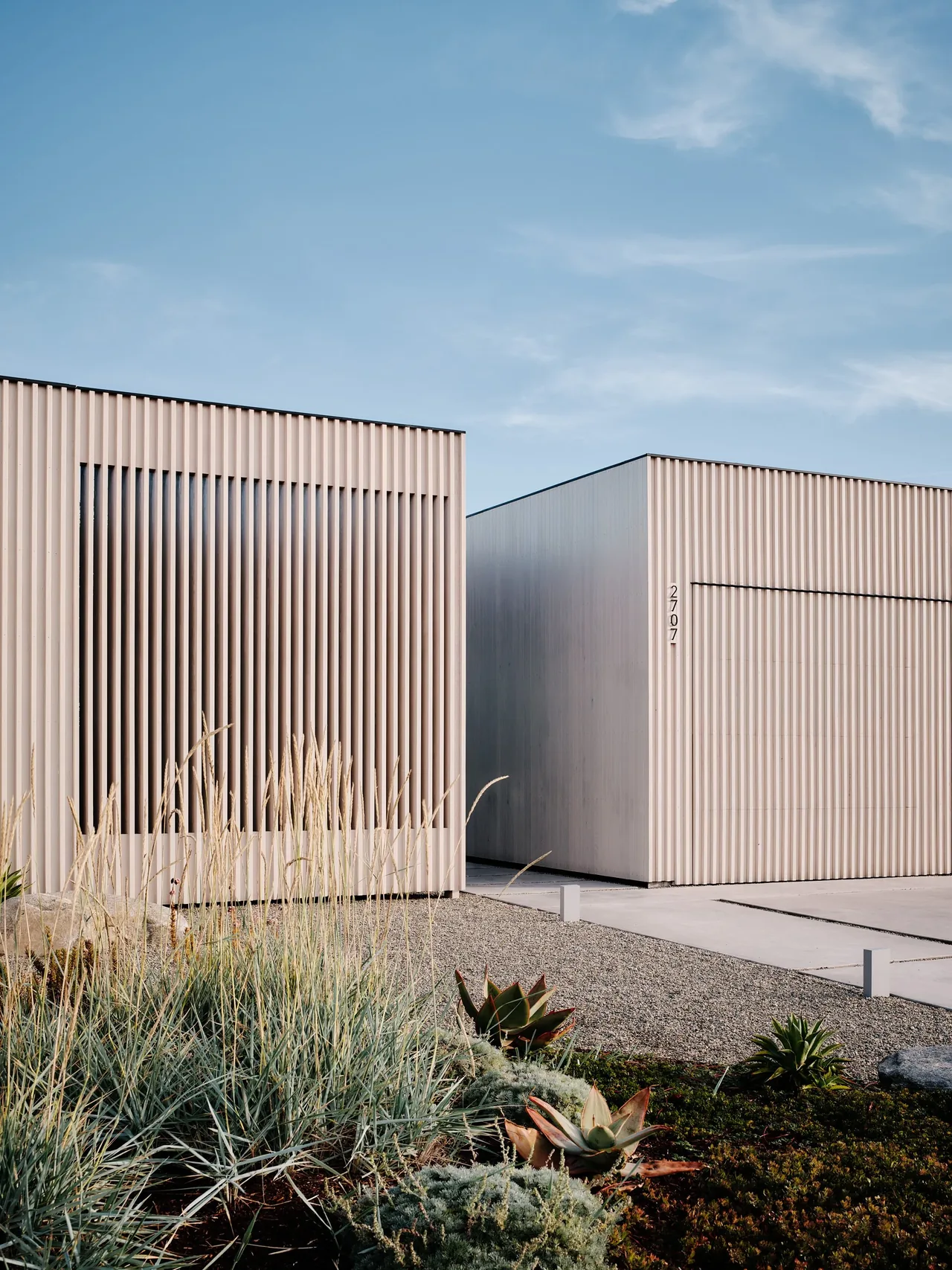 Carmel Meadows features two structures with vertical wood siding and a gravel driveway, framed by drought-tolerant landscaping.