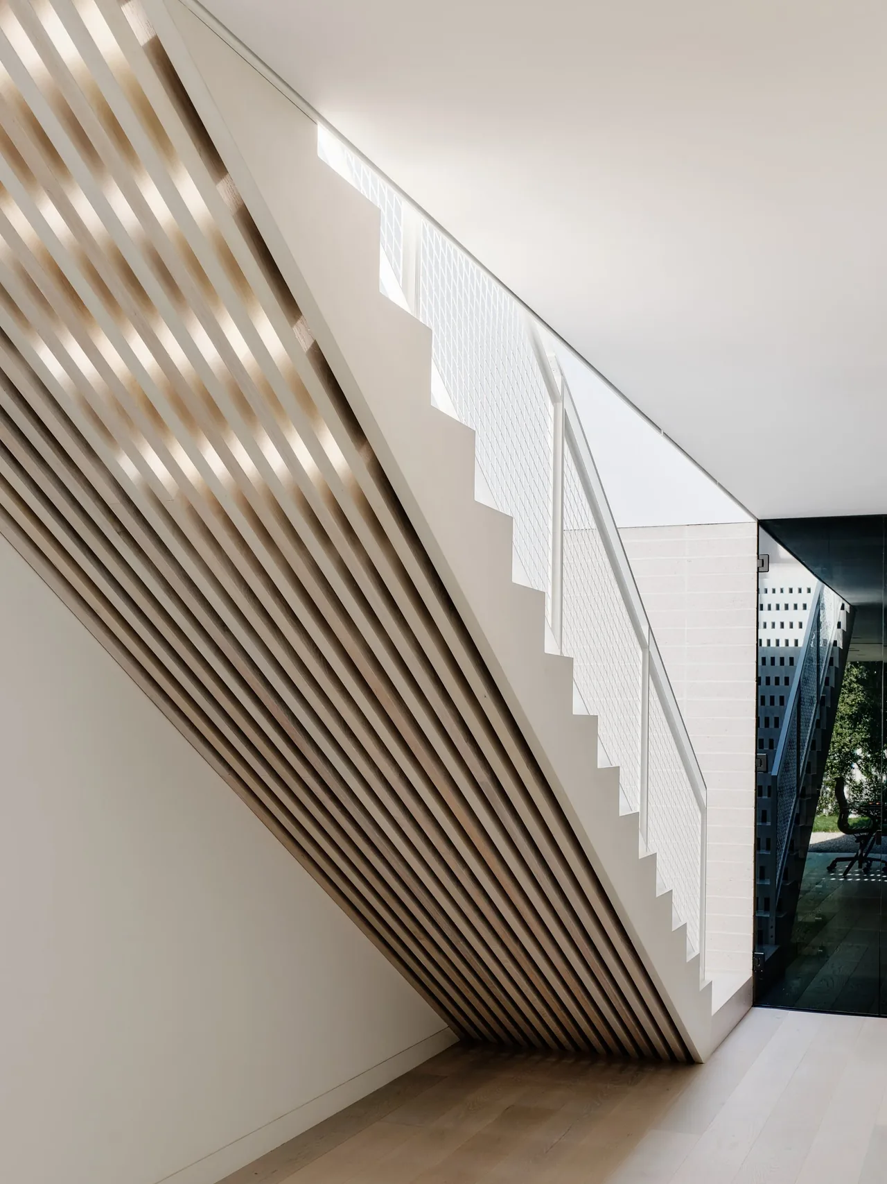 A white modern staircase with vertical wood slats underneath and metal mesh railing beside a glass door leading outside.