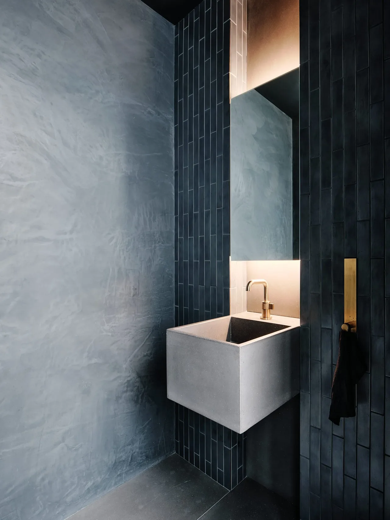 Compact bathroom sink with concrete basin, brass faucet, and dark vertical tiles backing a lit mirror niche.