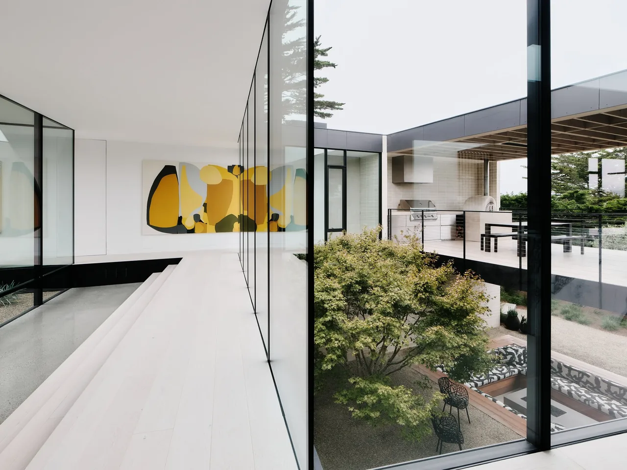 Modern home interior hallway with floor-to-ceiling glass walls overlooking a courtyard with a tree and outdoor seating area.