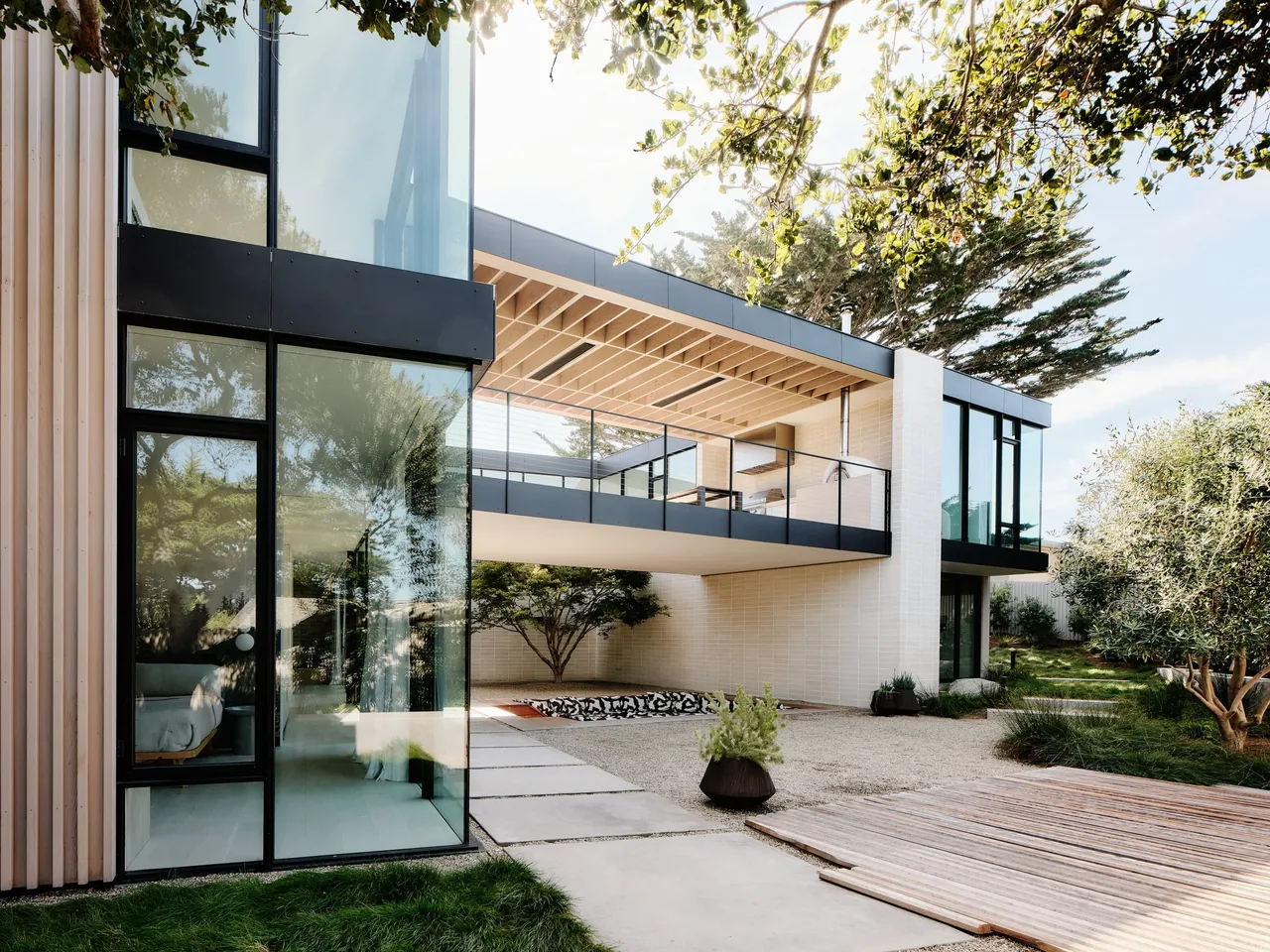 The Carmel Meadows project features a minimalist courtyard with vertical wood siding and expansive reflective glazing surrounded by gravel ground and landscaping.