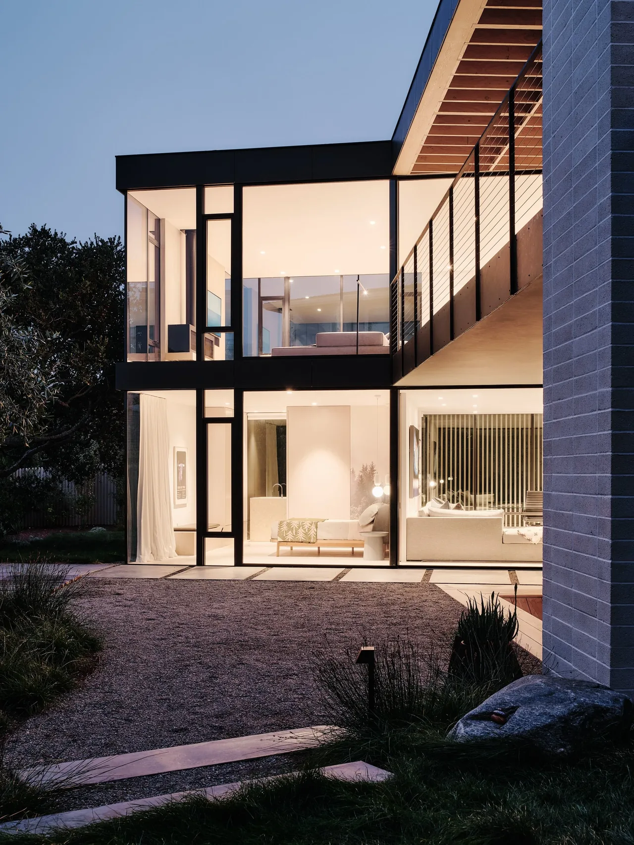 The Carmel Meadows house features expansive floor-to-ceiling windows and a cantilevered balcony on the upper level, showcasing indoor living spaces.