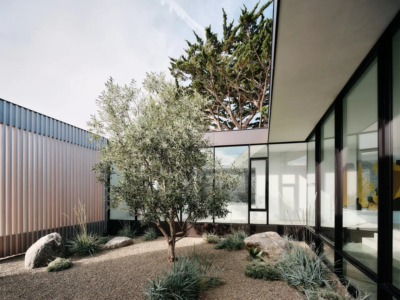 Modern courtyard featuring gravel landscaping, a solitary tree, vertical metal mesh railing, and large glass window panels.