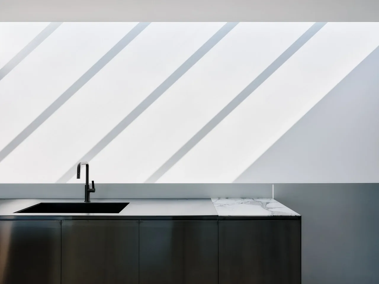 Modern kitchen counter features a black sink and faucet beneath white walls and ceiling with angled shadows.