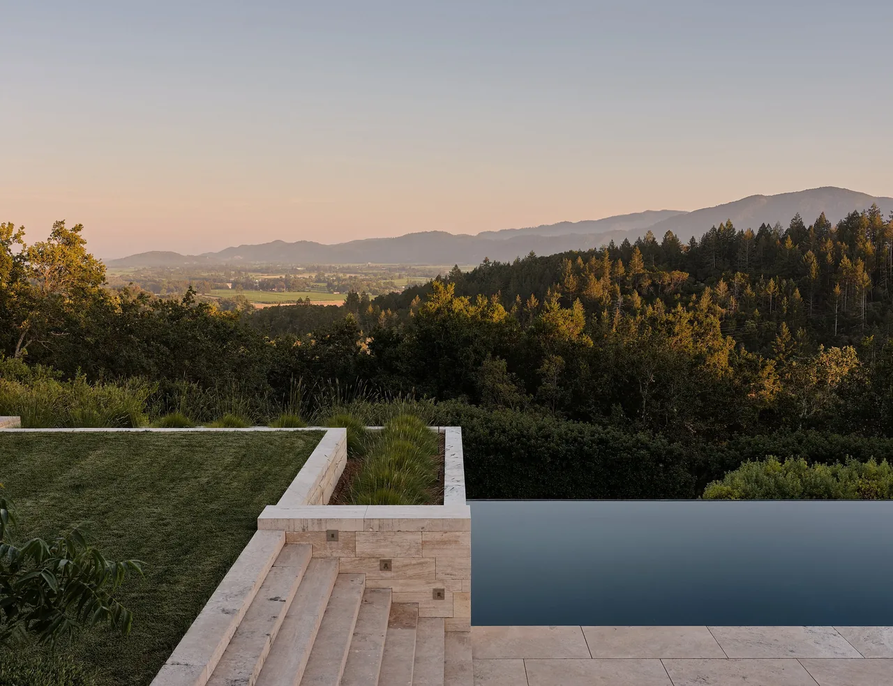 Infinity pool and stone steps overlook a forested valley and distant hills at sunset in the St Helena project.