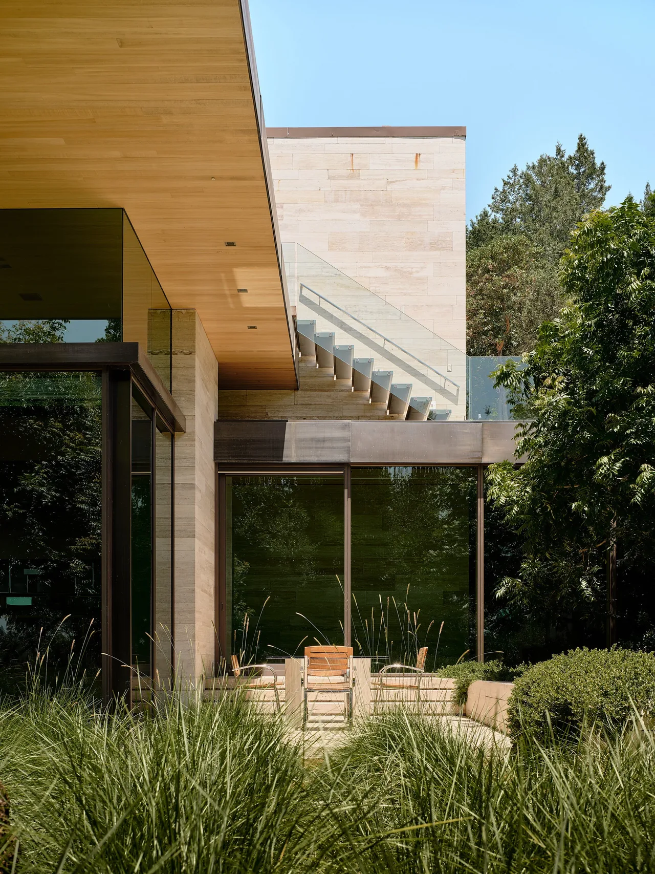 Exterior patio seating area with built-in benches and a rectangular table, next to a modern home with large glass panels and wood soffit.