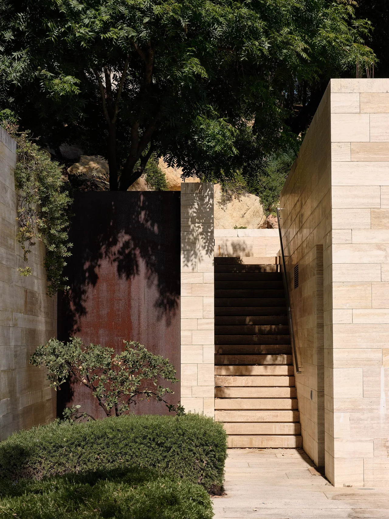 Exterior stone steps lead up between two light stone walls beside manicured shrubbery and a textured dark metal panel.