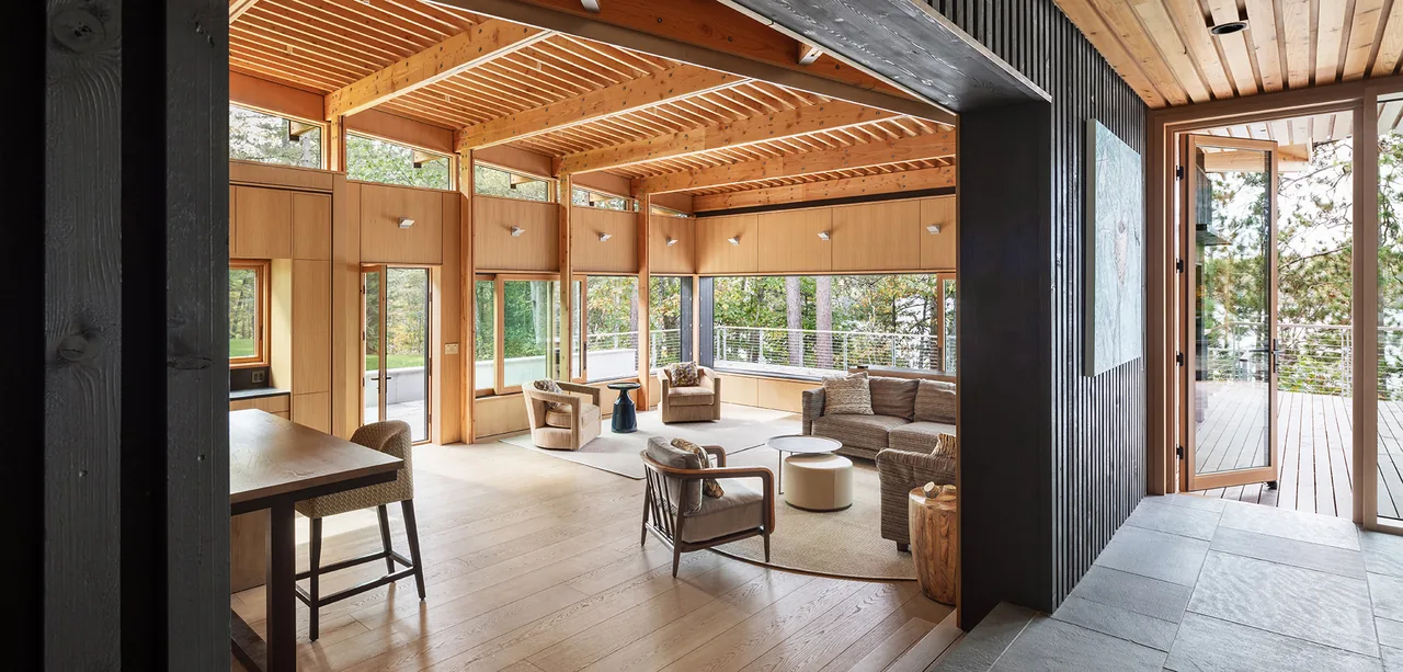 Living room with wood panel ceiling, large wood-framed windows, and access to an outdoor deck overlooking a forest.