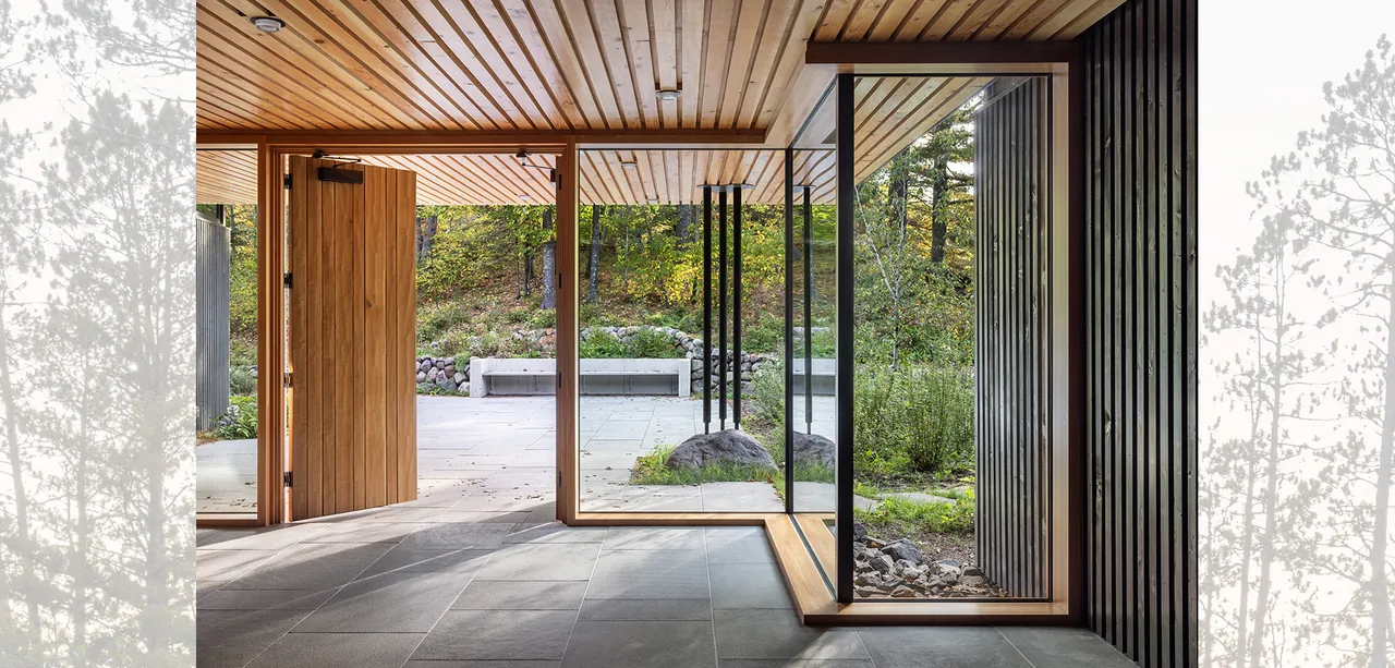 The Nest features a wood-paneled ceiling, large glass walls, and a pivot wooden entrance door opening to a stone patio with bench seating