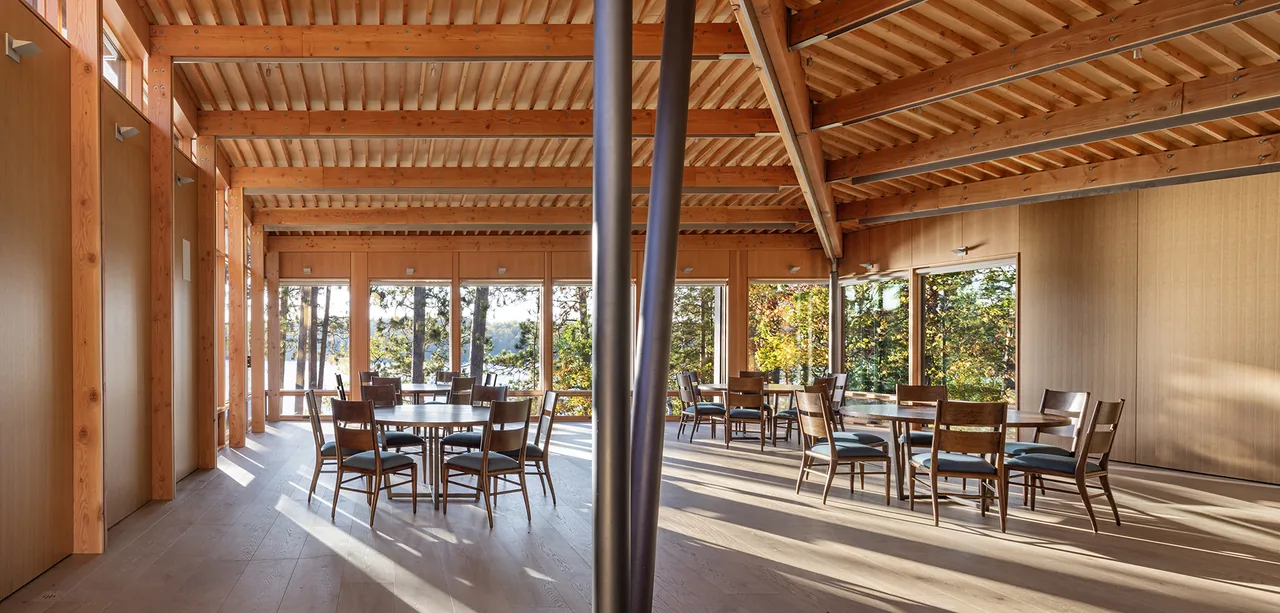Dining area with exposed wooden beams and large windows overlooking a natural wooded landscape.