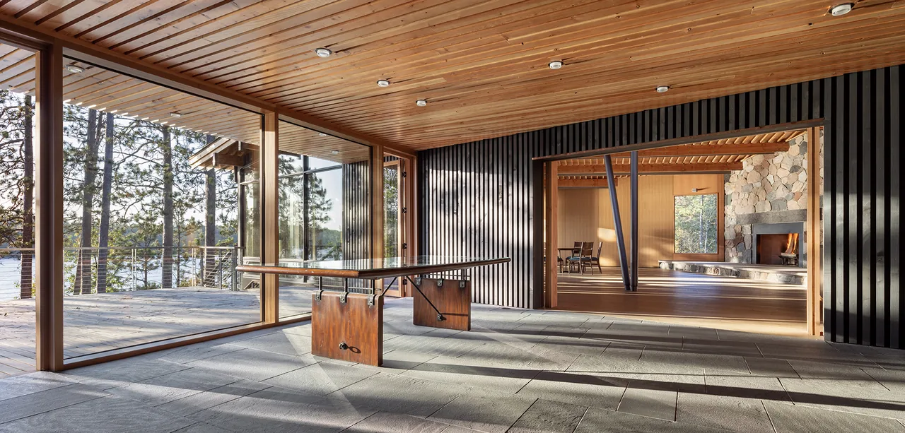 Covered patio with wood ceiling and black vertical siding opens to living room with stone fireplace at The Nest by SALA Architects.