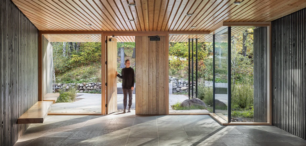 Entry area showing the large wood panel front door, black vertical siding, wood panel ceiling, and floor-to-ceiling glazing to natural landscape.
