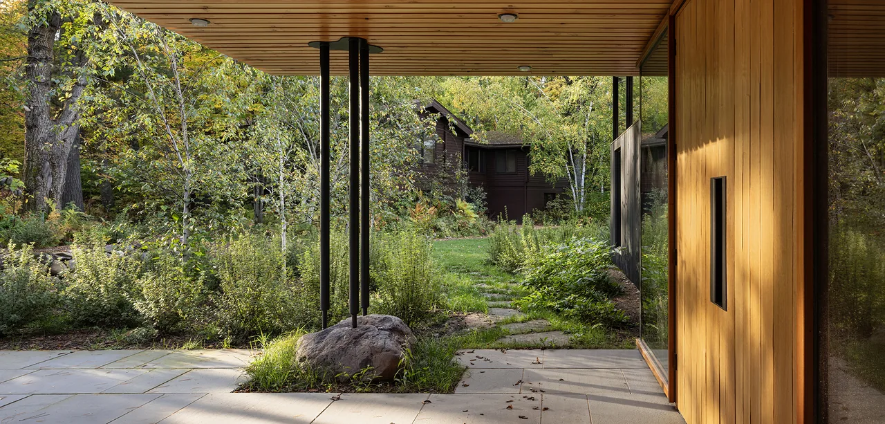 The Nest features a covered patio with a wood panel ceiling and steel columns framing views of a garden path and surrounding trees.