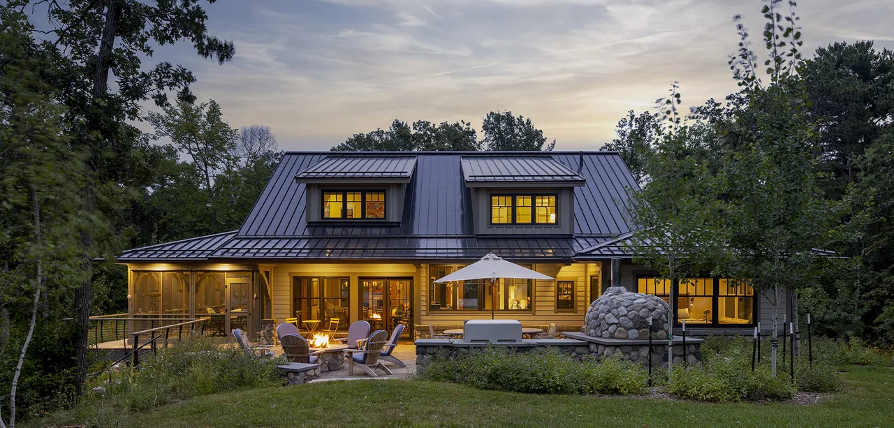 Tall Tree Lodge rear exterior shows metal shed roof, dormers, stone oven, and warm light through multiple windows.