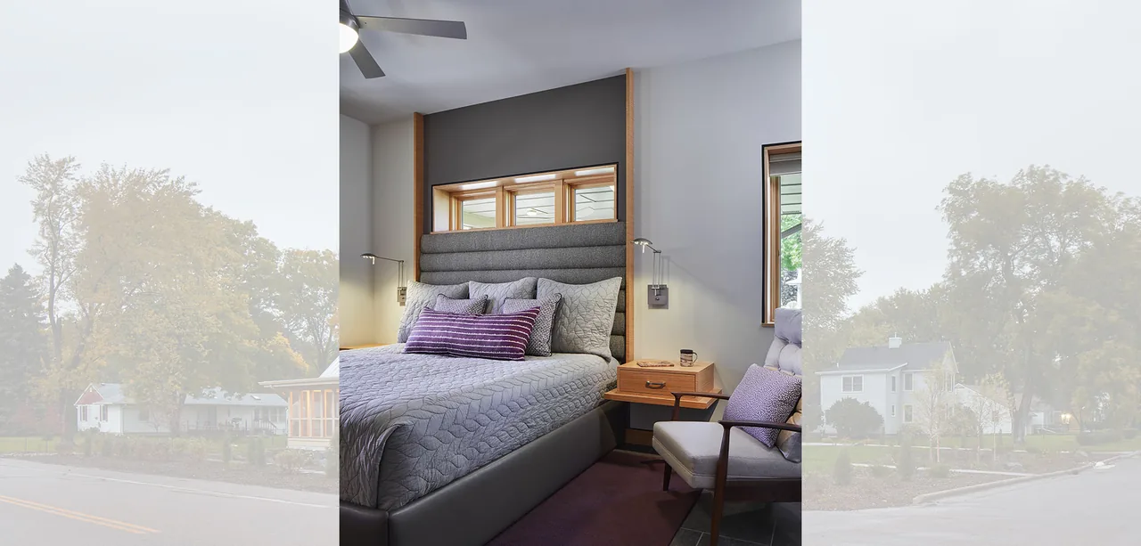 A bedroom features a gray upholstered bed with purple and gray pillows, a wooden nightstand, and small horizontal upper window for natural light.