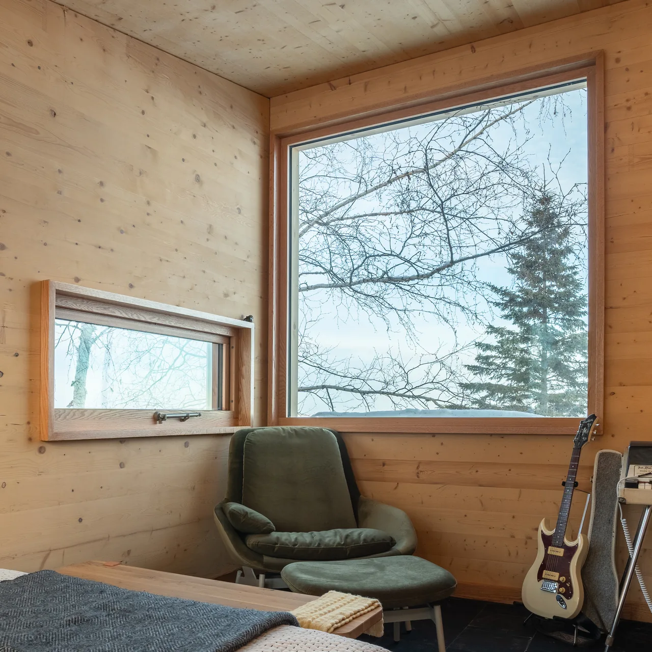 A wood-paneled room features large picture windows framing a wintry landscape with bare trees and an evergreen.