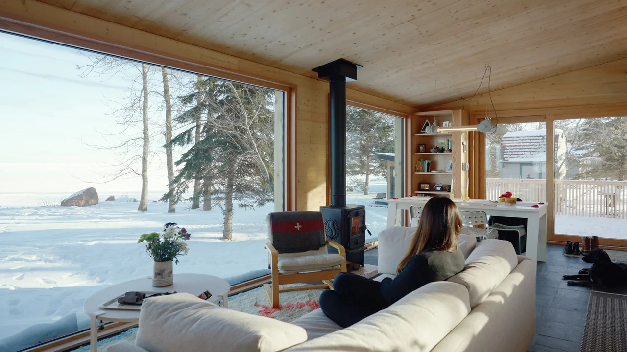 Living room with wood-paneled ceiling and walls, large wood-framed windows, and a wood stove overlooking snow-covered yard.