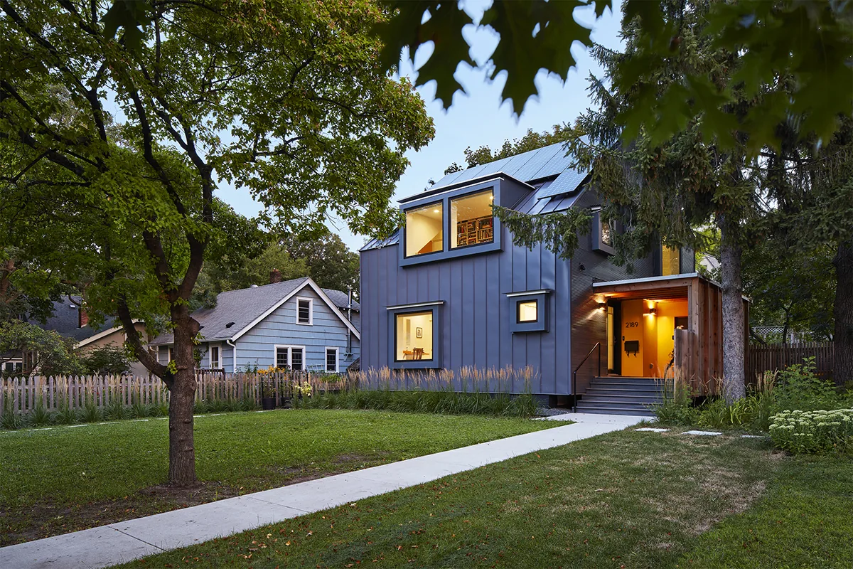 The Electric Bungalow features black metal siding, flat window frames, solar panels on the roof, and a richly planted front yard.