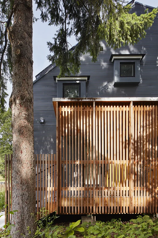 The Electric Bungalow features dark metal siding with flat window frames and a vertical wood slatted pergola covering part of the porch area.