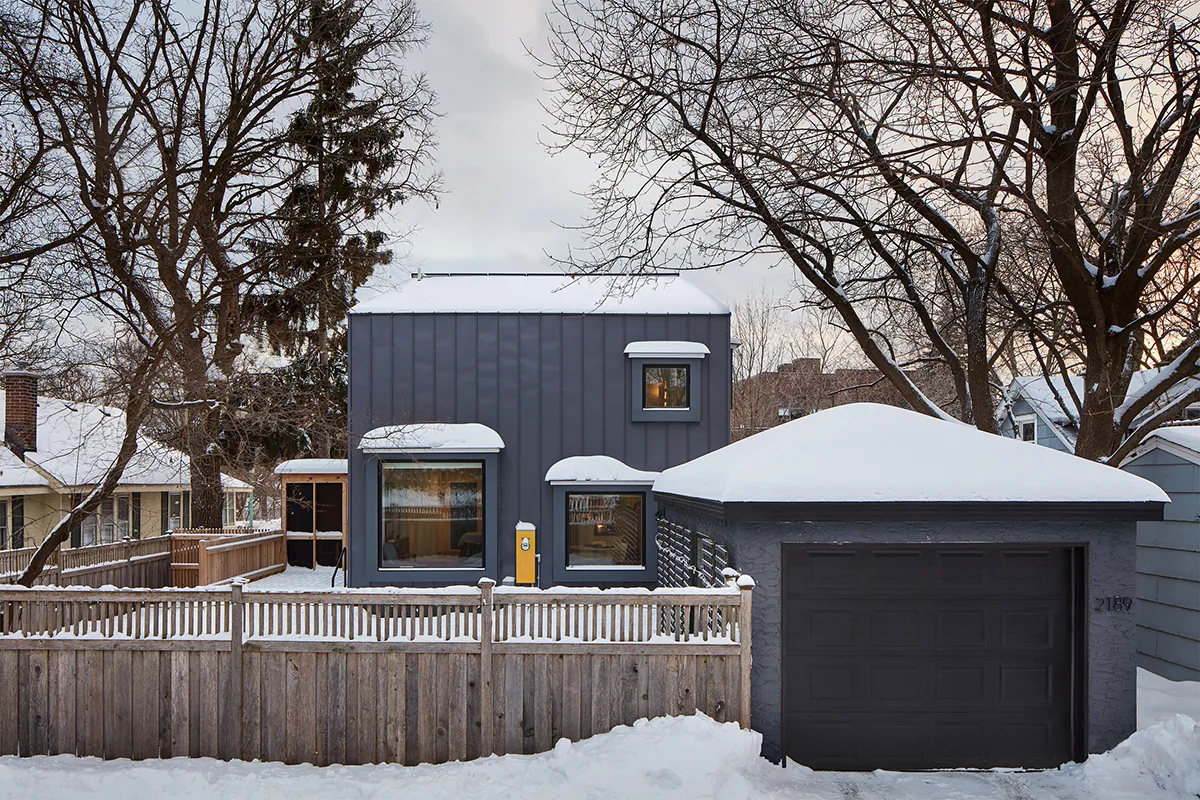 Modern dark gray house with corrugated metal siding and a separate garage, surrounded by a wooden fence in a snowy setting.