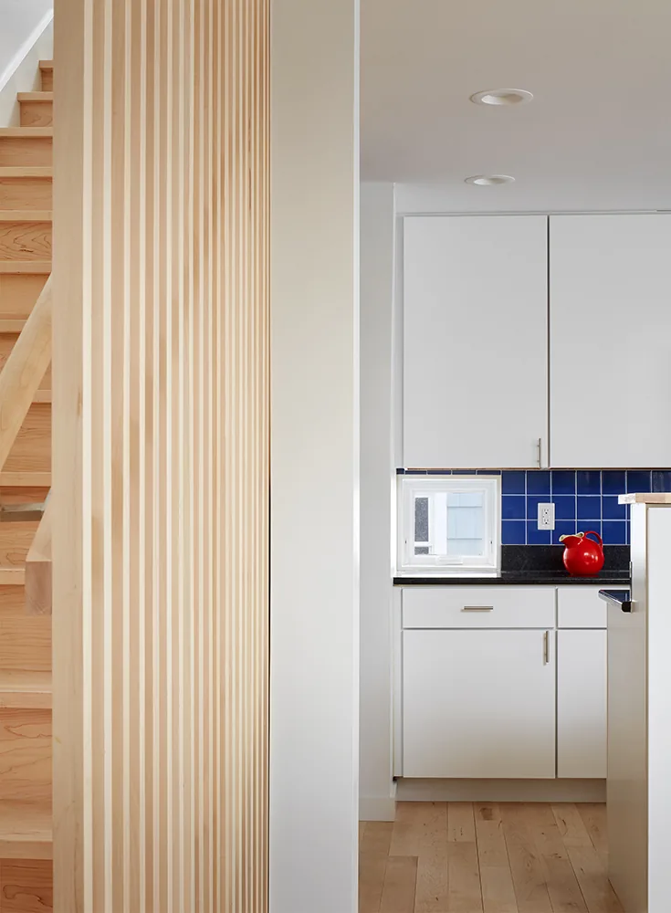 Kitchen features white cabinetry, black countertop, and blue tile backsplash beside a vertical wood slat staircase.