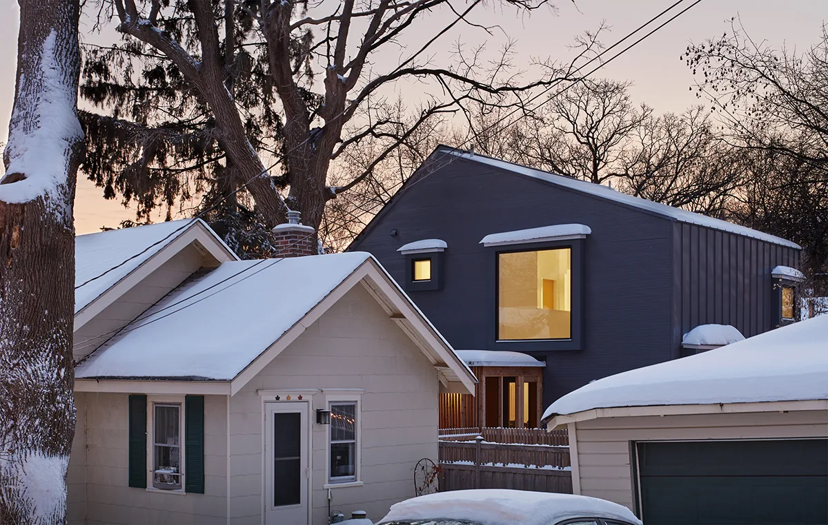 A modern dark gray house with square windows stands behind older snow-covered houses and a garage.
