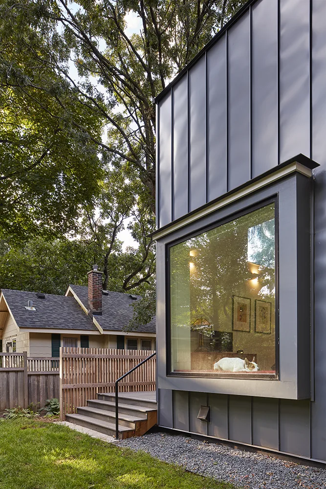 Modern home exterior with standing seam metal siding and a large window showing an interior with wall art and lighting.
