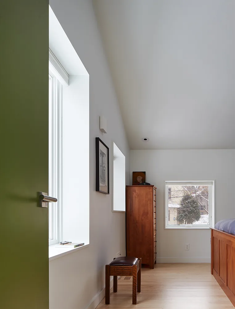 Bedroom with slanted ceiling and green door featuring a wooden dresser and small bench near windows.