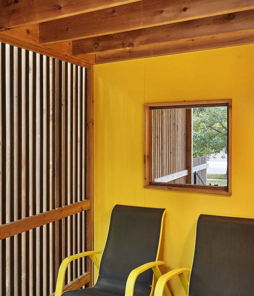 Outdoor seating area features black chairs with yellow armrests beneath a wooden pergola and a yellow painted wall with a small window.