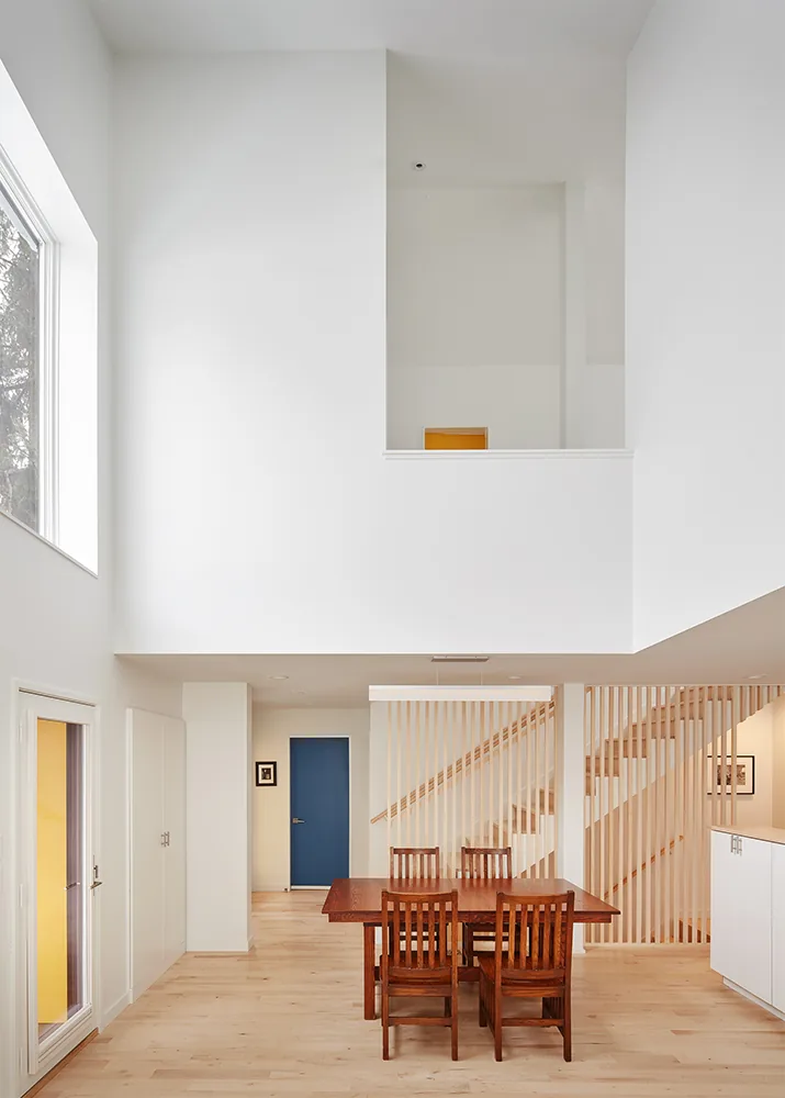 Dining area features a wooden table with four chairs and a staircase with vertical slats in a white, bright space.