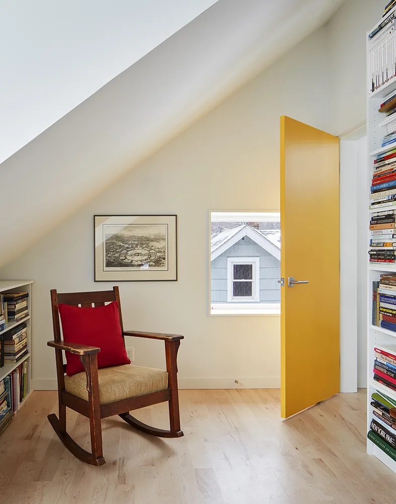 This narrow upstairs room has a wooden rocking chair, open yellow door, and bookshelves under a sloped ceiling.
