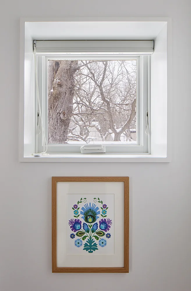 Square window with white frame and blind over floral framed art on interior gray wall.
