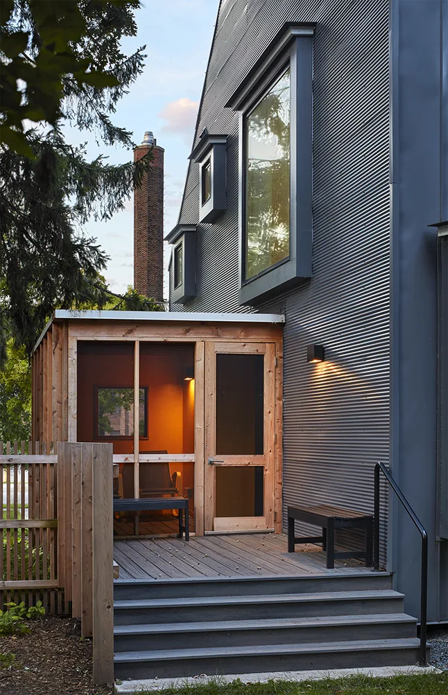 Rear exterior view of a modern home with dark metal siding and a wood-framed screened porch on a wooden deck accessed by stairs.