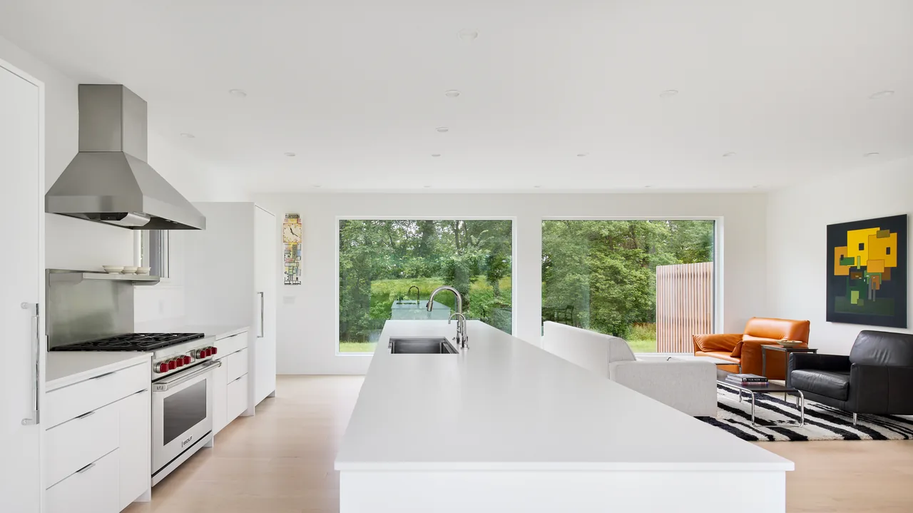 Open kitchen and living room with large windows framing a green outdoor view, featuring a long kitchen island and modern furnishings.