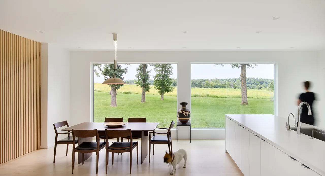 Minimalist dining and kitchen space with large black-framed rectangular windows facing a green field.