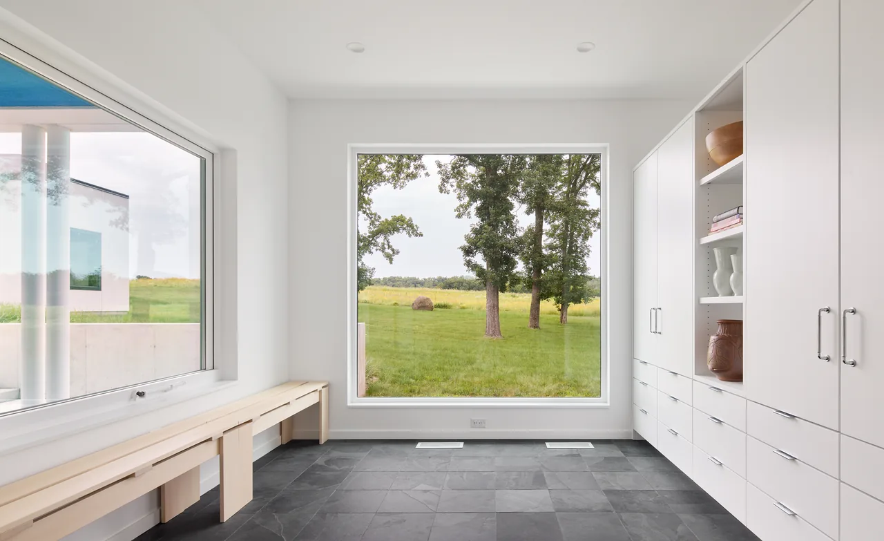 Bauhaus Farm minimalist room features a large square picture window framing the grassy field outside with white built-in cabinetry inside.