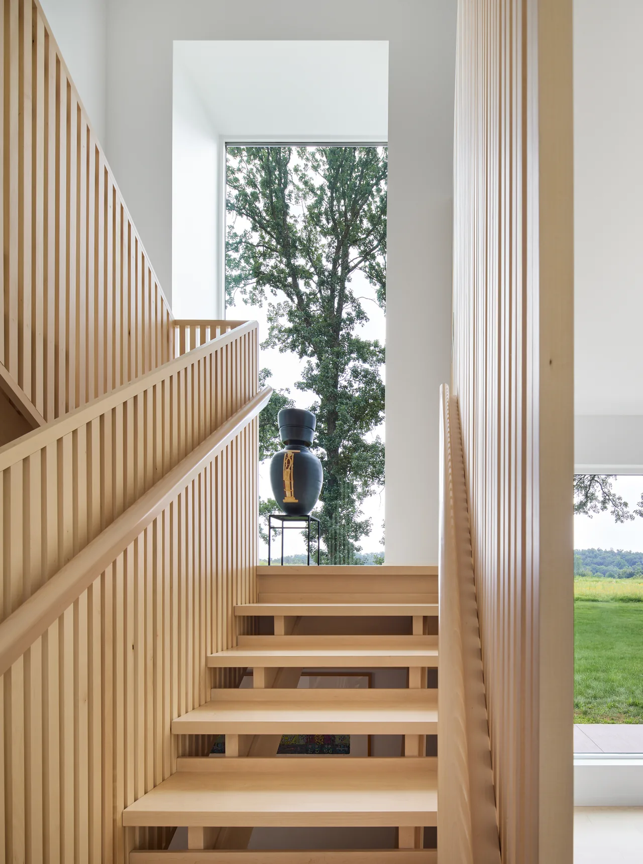A light wood staircase features vertical slat walls and a large window framing an outdoor tree.
