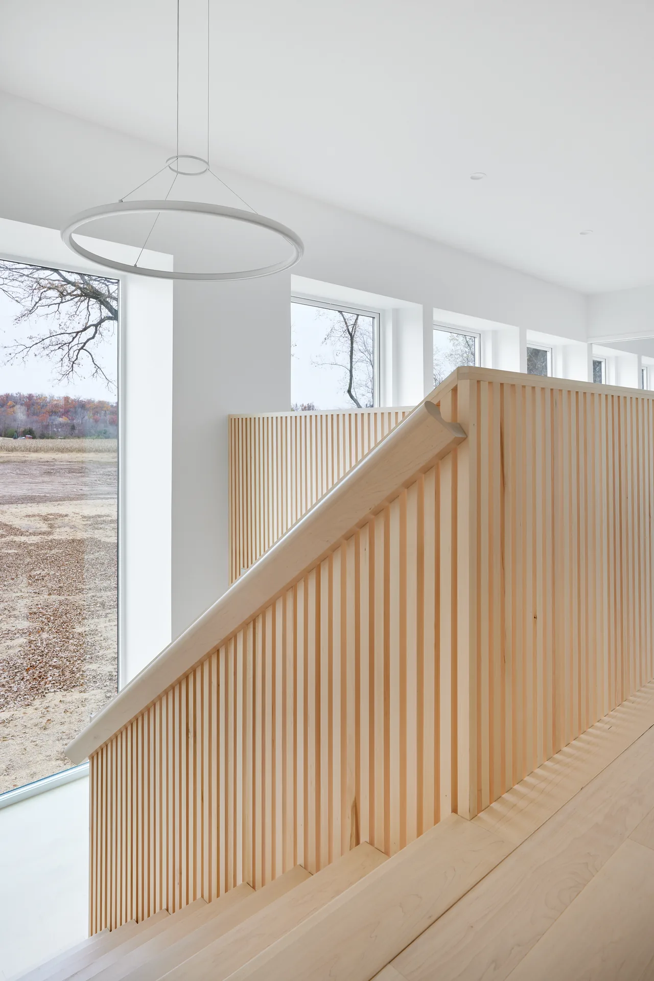 A light wood staircase with vertical slatted railing and a circular pendant light near large windows.