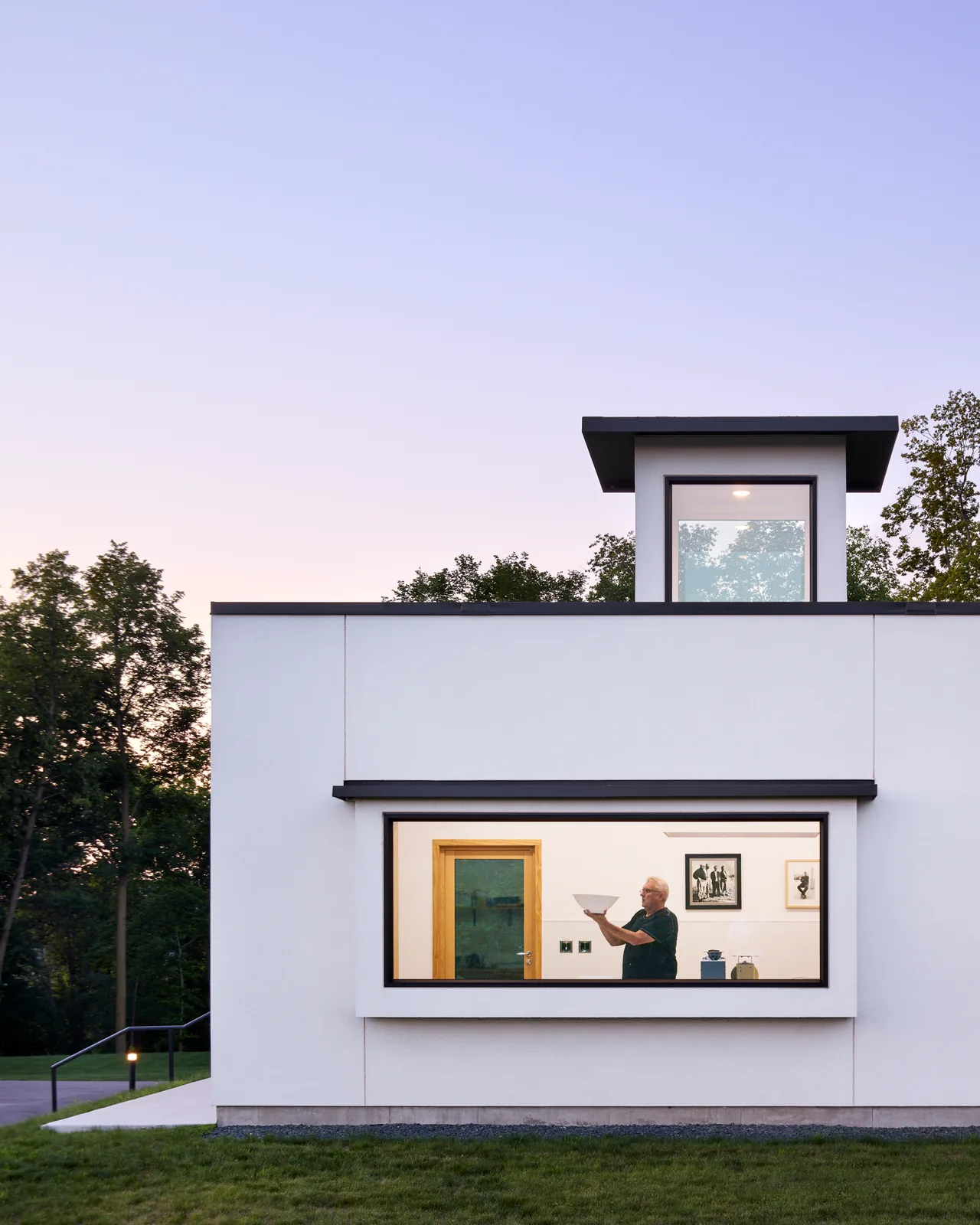 Minimalist white cubic volume farmhouse at dusk with large horizontal window and a single man inside.