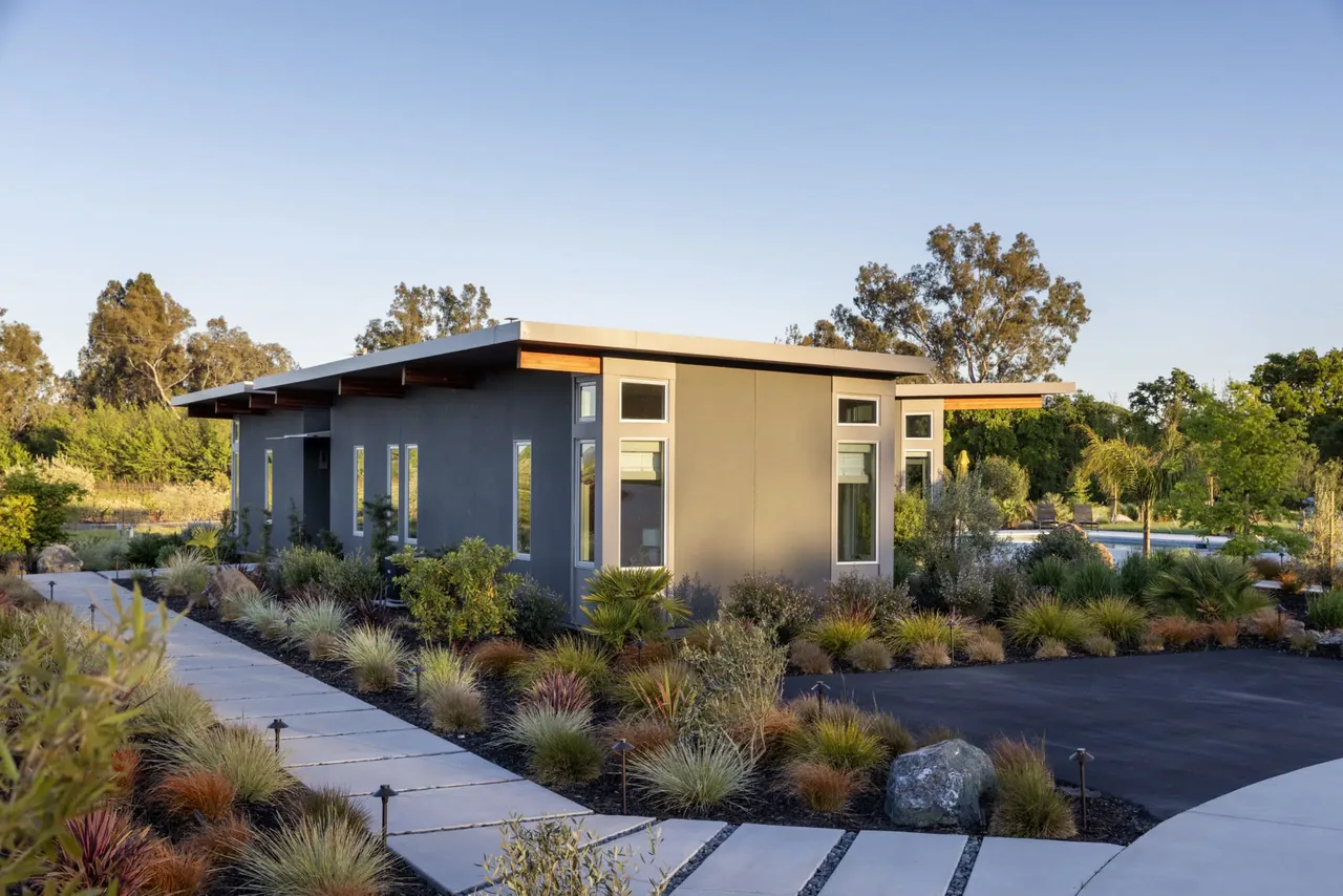 Modern home exterior with a flat roof and wood ceiling planks, featuring landscaped garden beds filled with ornamental grasses and large garden rocks.