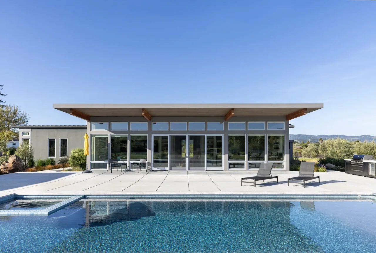 A modern flat-roof house with extensive floor-to-ceiling glazing overlooking a swimming pool in a rural setting.