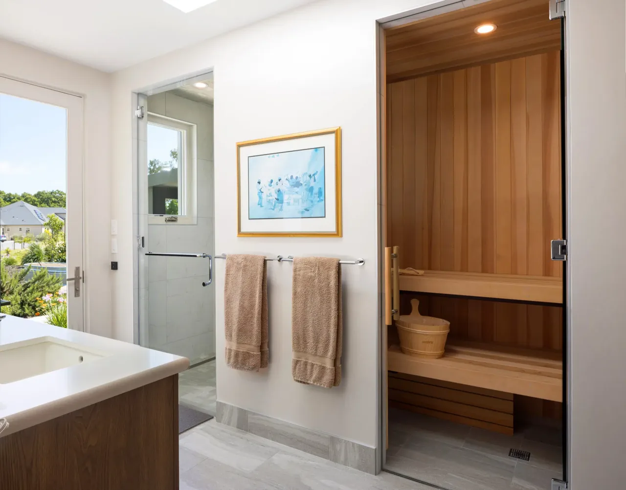 Bathroom interior featuring a wooden sauna, beige towels, and natural light from a window and glass door.