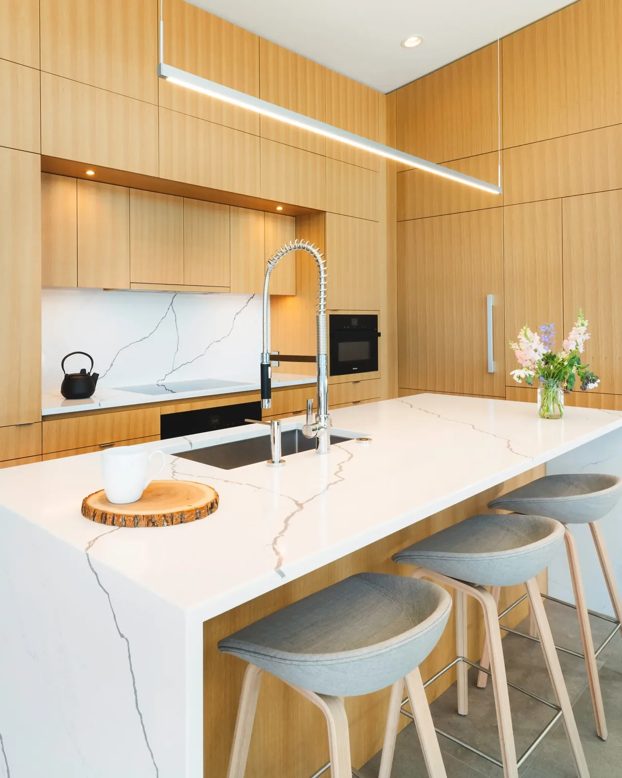 Kitchen with light wood cabinets, white marble island, built-in appliances, and suspended linear lighting.